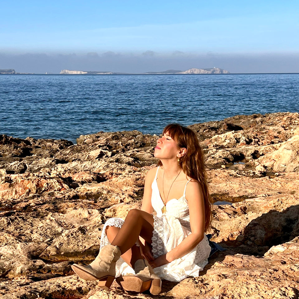 Hübsche Frau in weissen Spitzenkleid und Goldschmuck auf Felsen auf Ibiza.